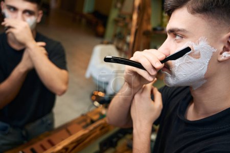 Foto de Joven con espuma en la cara afeitándose mientras mira el espejo en la barbería - Imagen libre de derechos