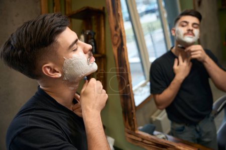 Foto de Hombre guapo peluquero afeitarse mientras mira el espejo en la barbería - Imagen libre de derechos