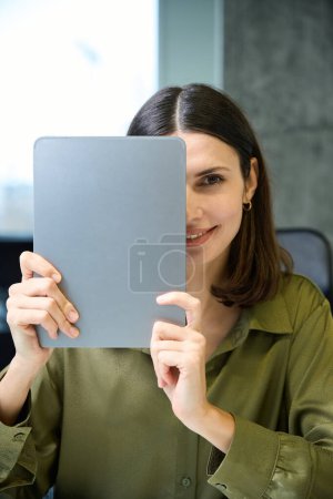 Téléchargez les photos : Heureuse femme d'affaires souriante couvrant visage avec tablette dans le bureau de l'entreprise - en image libre de droit