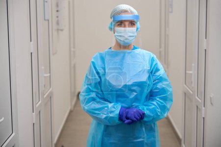 Photo for Female infectious disease specialist stands in a bright corridor in protective equipment, she uses a protective shield - Royalty Free Image