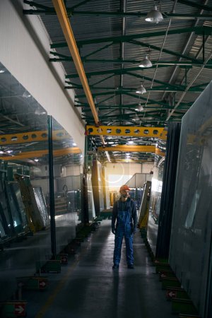 Foto de El hombre con overol está parado en un taller de producción de ventanas, un trabajador con un casco de seguridad naranja - Imagen libre de derechos