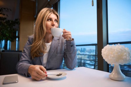 Téléchargez les photos : Femme blonde en costume d'affaires profite d'une tasse de café dans un restaurant confortable, avec un paysage urbain devant la fenêtre - en image libre de droit