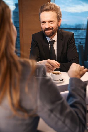 Téléchargez les photos : Élégant homme d'affaires bavarde avec une femme autour d'une tasse de café, paysage urbain devant la fenêtre - en image libre de droit