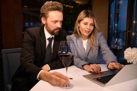 Foto de La gente en trajes de negocios se sienta en un restaurante con una computadora portátil, vasos de vino en la mesa - Imagen libre de derechos
