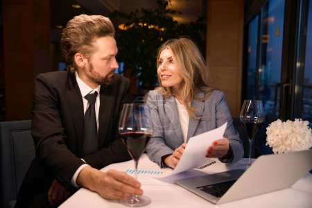 Foto de Hombre y una mujer tomando una copa de vino en un restaurante de moda, documentos y una computadora portátil sobre la mesa - Imagen libre de derechos