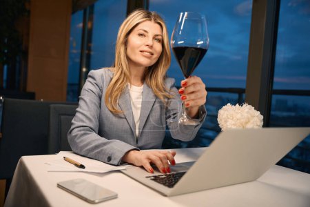 Foto de Lady en un traje de negocios disfruta de una copa de vino en el trabajo, un paisaje urbano fuera de la ventana del restaurante - Imagen libre de derechos
