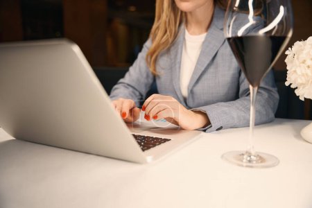 Téléchargez les photos : Femme seule dans la salle de restaurant avec un ordinateur portable et un verre de vin, elle est dans un élégant costume d'affaires - en image libre de droit