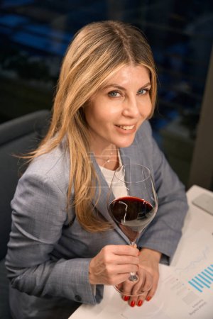 Téléchargez les photos : Dame souriante appréciant le vin rouge, elle porte un élégant costume d'affaires - en image libre de droit