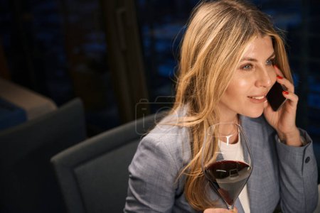 Foto de Señora sonriente se está comunicando en su teléfono móvil y disfrutando del vino tinto, ella está usando un elegante traje de negocios - Imagen libre de derechos