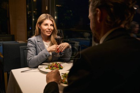 Foto de Empresaria de mediana edad se comunica con un hombre durante la cena en un restaurante, hay vino tinto en su copa - Imagen libre de derechos
