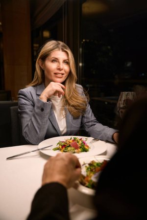 Téléchargez les photos : Belle femme d'affaires dînant dans un restaurant confortable avec un partenaire d'affaires, salade légère sur des assiettes - en image libre de droit