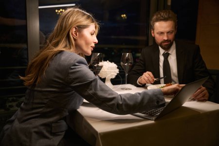 Téléchargez les photos : Femme d'affaires élégante dînant dans un restaurant confortable avec un partenaire d'affaires, ordinateur portable sur la table - en image libre de droit