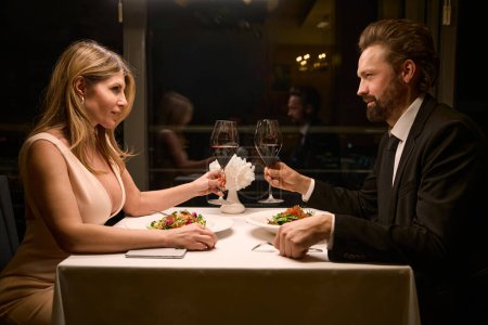 Foto de Mujer en un vestido de noche y hombre están hablando durante la cena en el acogedor restaurante, un hombre en un elegante traje de negocios - Imagen libre de derechos