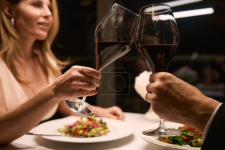 Téléchargez les photos : Femme en robe de soirée et son compagnon célèbrent dans un restaurant confortable, il y a du vin rouge dans les verres - en image libre de droit