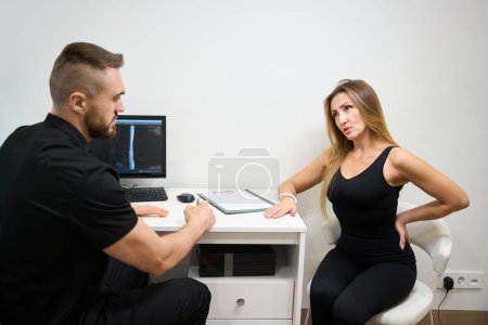 Téléchargez les photos : Physiothérapeute mène une consultation dans un bureau lumineux, le patient se plaint de maux de dos - en image libre de droit