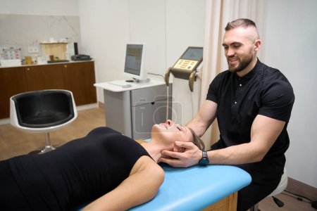 Spezialist massiert während einer manuellen Therapiesitzung eine Frau im Nacken, die Patientin liegt auf dem Massagetisch