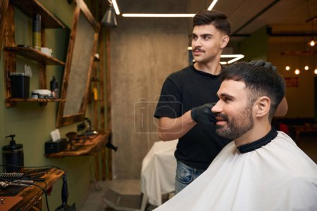 Téléchargez les photos : Homme barbu se faire couper les cheveux par coiffeur tout en étant assis sur la chaise au salon de coiffure - en image libre de droit