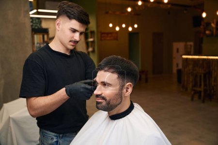 Téléchargez les photos : Barber en utilisant une tondeuse à cheveux pour faire une coupe de cheveux élégante pour le client au salon de coiffure - en image libre de droit