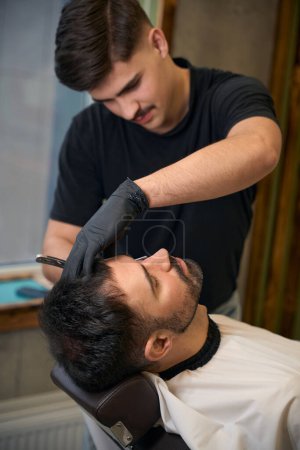 Photo for Bearded man getting shaved with straight edge razor by hairdresser at barbershop - Royalty Free Image