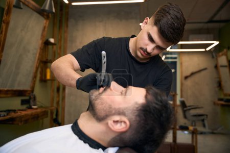 Téléchargez les photos : Homme barbu se faire couper les cheveux de barbe par le coiffeur tout en étant assis sur la chaise au salon de coiffure - en image libre de droit