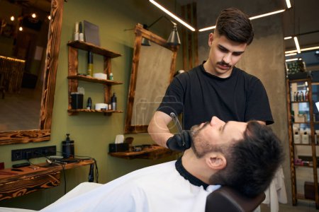 Foto de Barbudo hombre consiguiendo barba corte de pelo por barbero mientras está sentado en silla en la barbería - Imagen libre de derechos