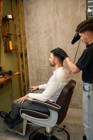 Téléchargez les photos : Jeune homme barbu se faire toiletter par un coiffeur avec sèche-cheveux au salon de coiffure - en image libre de droit