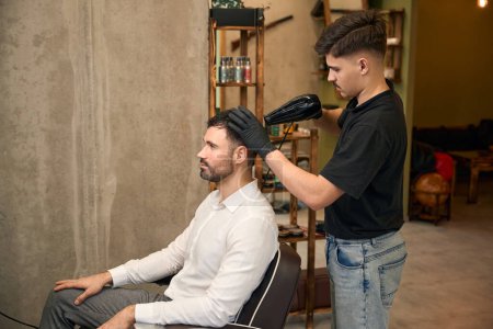 Photo for Hairdresser blow drying his clients hair in professional barbershop - Royalty Free Image