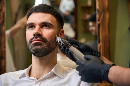 Téléchargez les photos : Homme barbu se faire couper la barbe avec tondeuse à cheveux par coiffeur au salon de coiffure - en image libre de droit