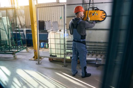 Foreman utiliza un dispositivo de elevación al vacío en un taller de producción, un hombre en mono de trabajo