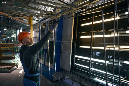 El capataz de mediana edad pega marcos espaciadores en una instalación de producción de ventanas, un hombre con un casco naranja