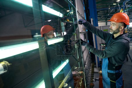 El hombre con casco de seguridad trabaja en un taller de producción produciendo ventanas modernas de doble acristalamiento