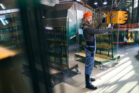 Foto de Hombre usando un dispositivo de elevación al vacío en una fábrica de ventanas, hombre en overoles - Imagen libre de derechos