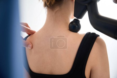 Téléchargez les photos : Physiothérapeute masse un dos de patients avec un masseur spécial, femme dans des vêtements confortables - en image libre de droit