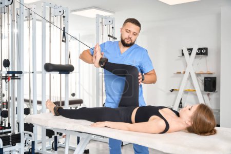 Foto de Fisioterapeuta experimentado utiliza una máquina de ejercicio especial para trabajar con un paciente, la mujer se acuesta en una mesa de masaje - Imagen libre de derechos