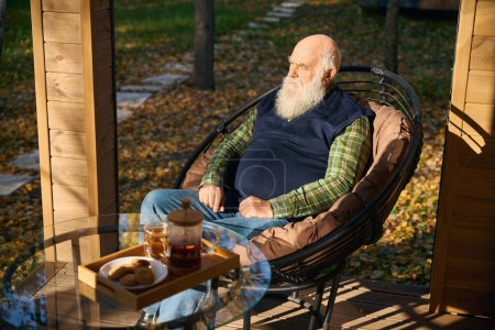 Téléchargez les photos : Vieil homme barbu prend le thé sur la véranda, il est assis dans une chaise confortable - en image libre de droit