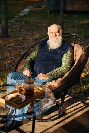 Téléchargez les photos : Le vieil homme aux cheveux gris prend le thé sur la véranda, il est assis sur une chaise confortable - en image libre de droit
