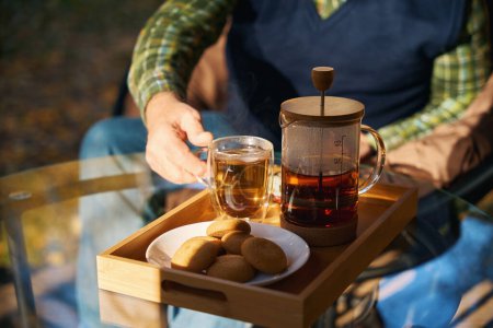 El hombre disfruta de la taza de té en la terraza de una casa de campo, hay una tetera y galletas en la mesa