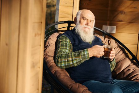 Grauhaariger alter Mann sitzt auf der Veranda mit einer Tasse Tee, er sitzt in einem bequemen Stuhl