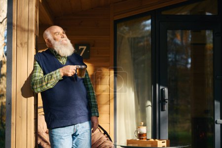 Foto de Anciano hombre de pelo gris disfruta de una taza de té en la terraza de una casa de campo, una tetera y galletas en la mesa - Imagen libre de derechos
