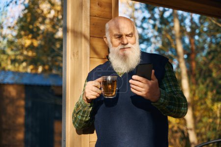 Téléchargez les photos : Vieil homme avec tasse de thé et téléphone portable sur la véranda, il y a beaucoup d'arbres près de la maison - en image libre de droit