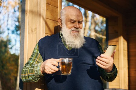 Téléchargez les photos : Vieil homme souriant avec un téléphone portable sur la véranda, il est dans des vêtements décontractés confortables - en image libre de droit