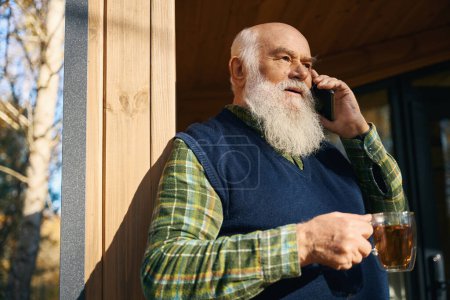 Téléchargez les photos : Homme âgé communique sur un téléphone portable sur la véranda, il a une tasse de thé dans la main - en image libre de droit