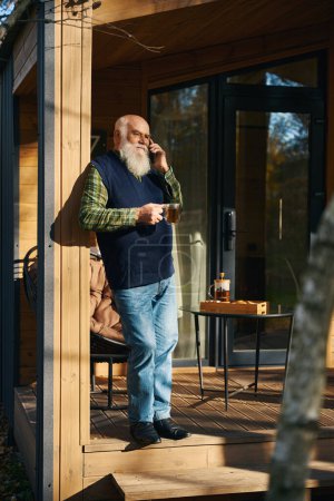 Téléchargez les photos : L'homme à barbe grise communique sur un téléphone portable sur la véranda, il a une tasse de thé à la main - en image libre de droit