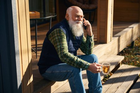 Téléchargez les photos : Le vieil homme à barbe grise se repose sur la véranda d'une maison de campagne, communiquant sur son téléphone portable - en image libre de droit