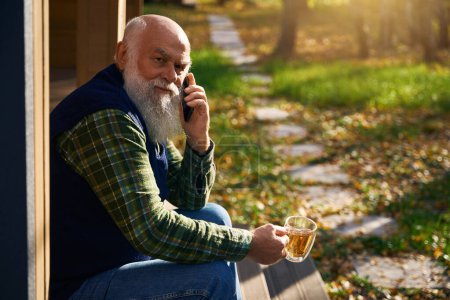 Foto de Viejo barbudo gris se relaja en la veranda de una casa de campo, se comunica por su teléfono móvil - Imagen libre de derechos