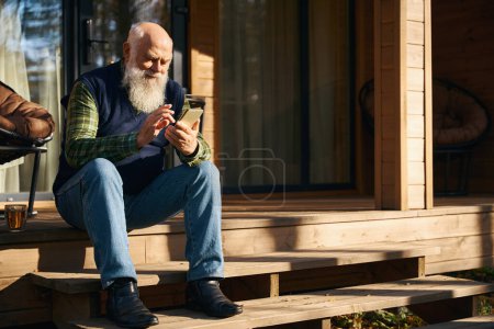 Téléchargez les photos : Le vieil homme est assis sur le porche d'une maison de campagne, il communique sur son téléphone portable - en image libre de droit