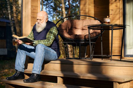 Téléchargez les photos : L'homme à barbe grise est assis avec un livre sur le porche d'une maison de campagne, il apprécie une tasse de thé - en image libre de droit