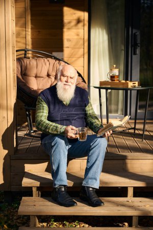 Téléchargez les photos : Le vieil homme est assis avec un livre sur le porche d'une maison de campagne, il apprécie une tasse de thé - en image libre de droit