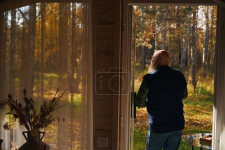 Foto de Hombre de barba gris admira el bosque de otoño, se encuentra en la terraza de una casa de campo - Imagen libre de derechos