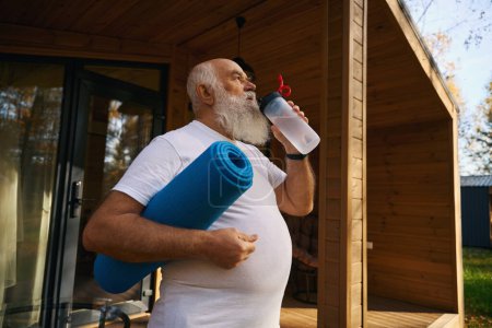 Foto de Fuerte anciano salió de la casa para los ejercicios de la mañana con una botella de agua y un karimat - Imagen libre de derechos